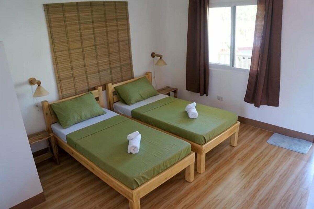 Two single beds with green bedding, wooden headboards, bedside tables with lamps, a window with curtains, and a wooden floor.