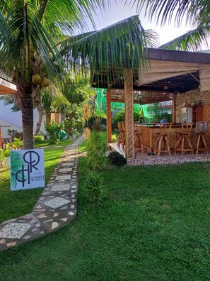 A tropical garden with a wooden bar and seating area, a sign for 'Hotel Renate', and a stone pathway.