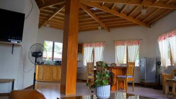 A spacious living area with a high ceiling, wooden beams, a central table with a plant, and a TV mounted on the wall.