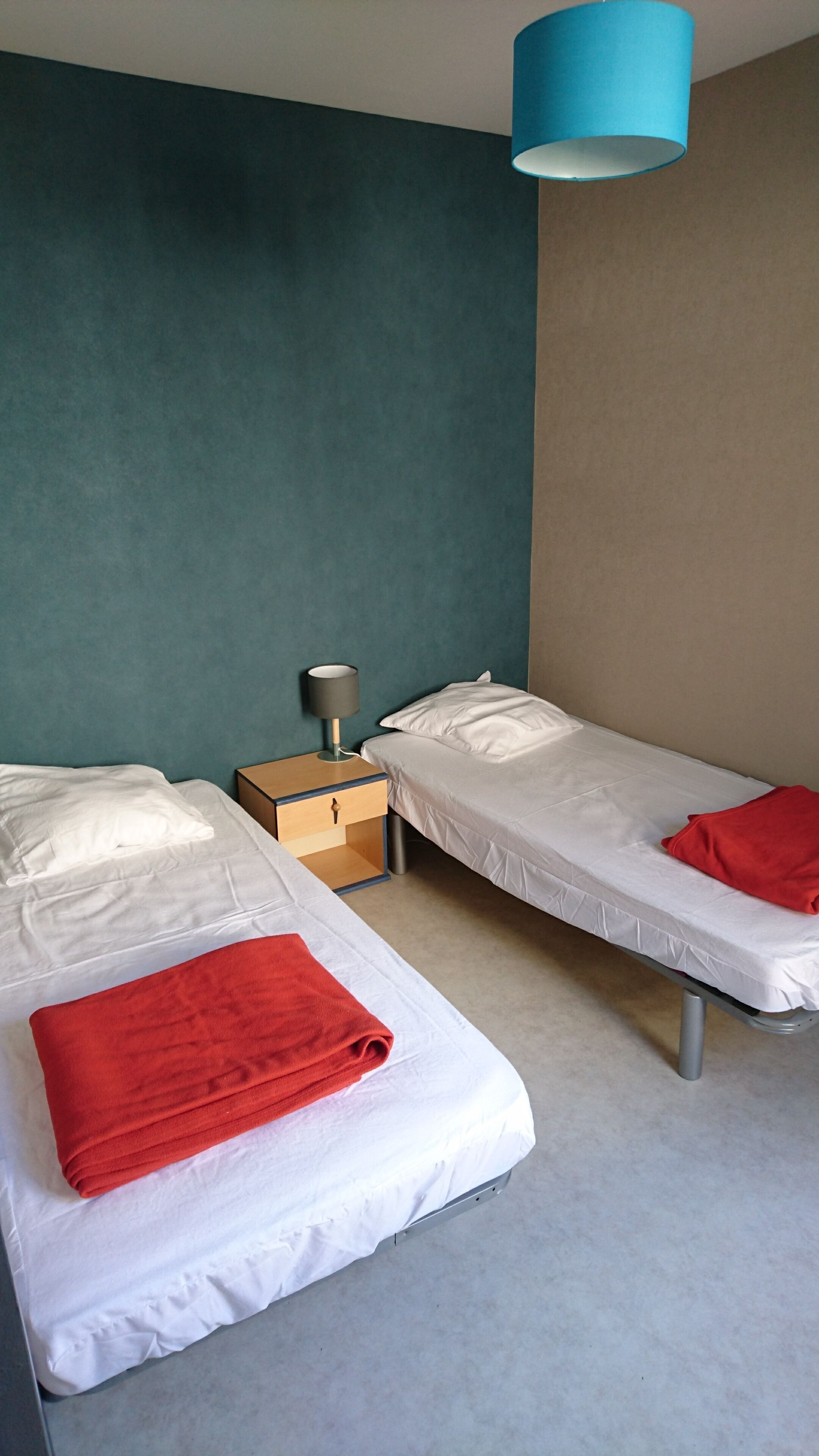 Una habitación con dos camas individuales, cada una con un colchón blanco y una manta roja. Entre las camas hay una mesita de noche de madera con una lámpara. Las paredes son color turquesa y beige, y hay un colgante de luz azul sobre las camas.