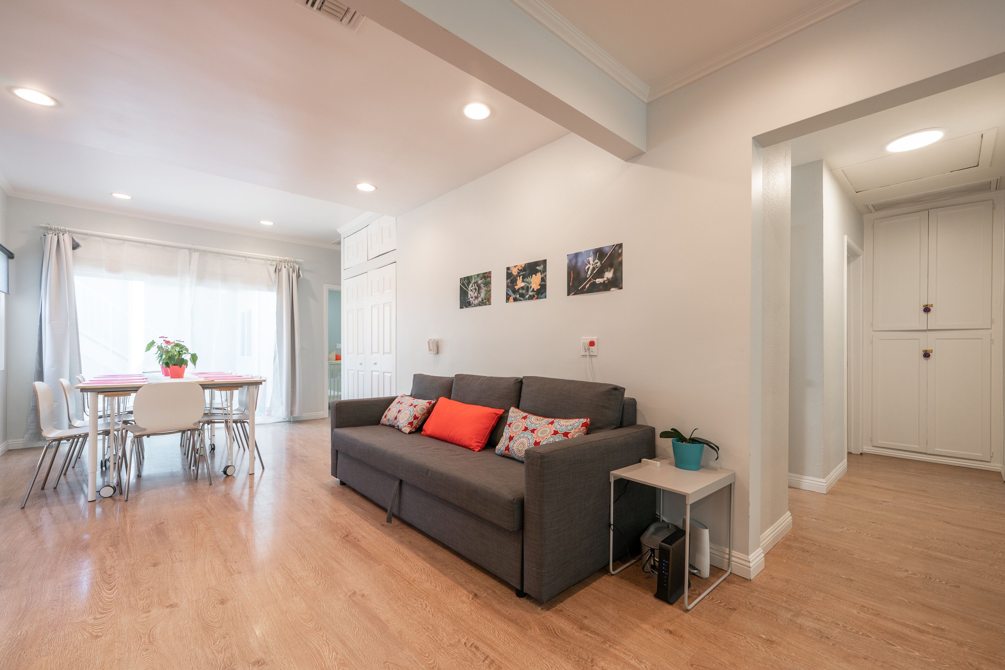 A modern living room with a grey sofa, a dining table, and a small side table. There are three wall-mounted artworks and a white door in the background.