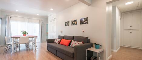 A modern living room with a grey sofa, a dining table, and a small side table. There are three wall-mounted artworks and a white door in the background.