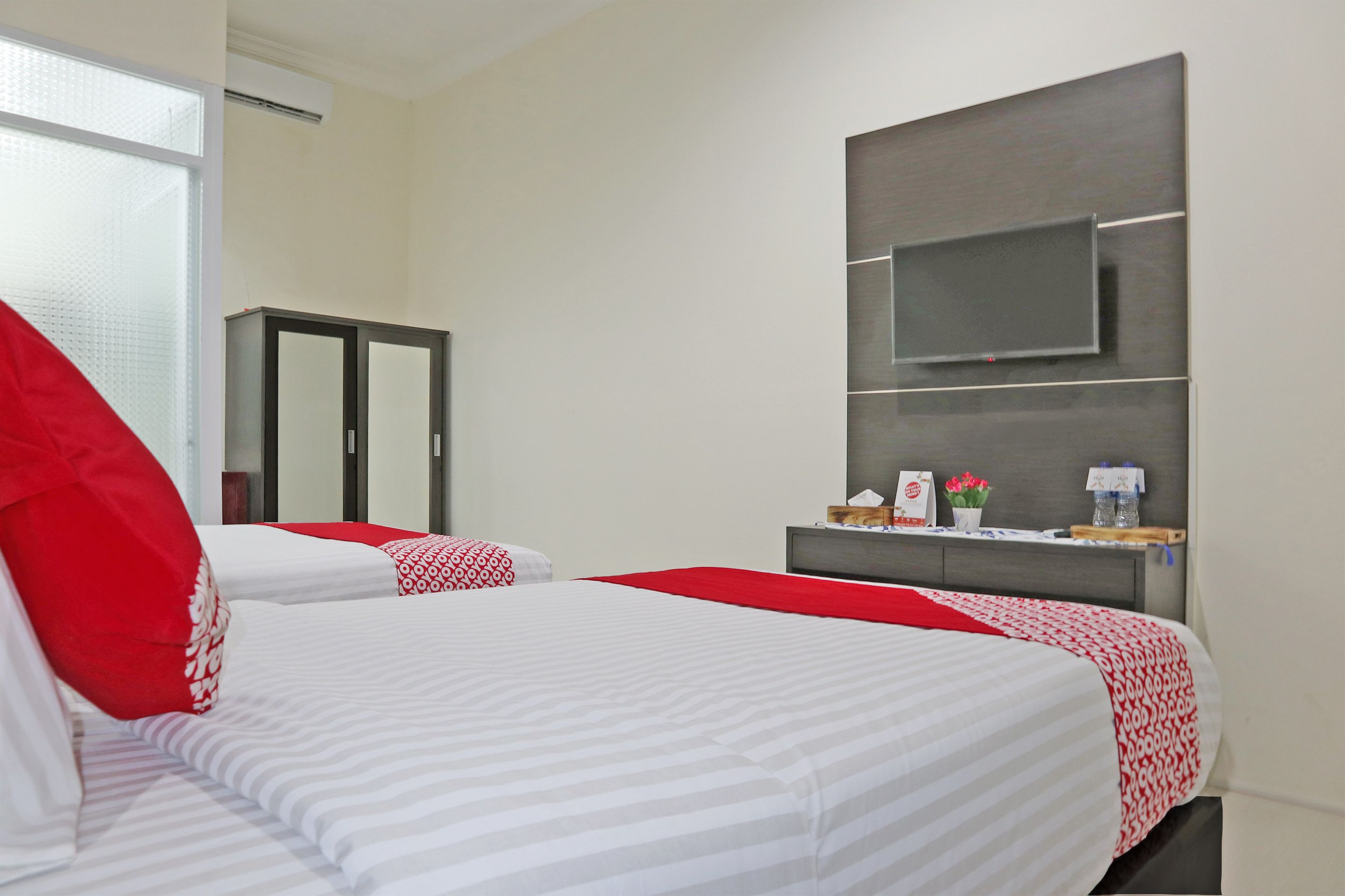 A hotel room with two beds, a TV mounted on the wall, a wardrobe, and a small table with a vase of flowers.