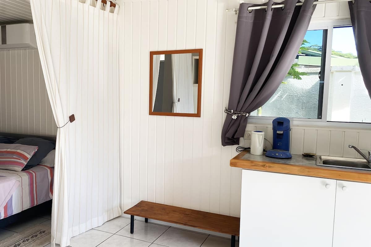 A bedroom with a bed, a bedside table with a kettle, a mirror, and a window with curtains.
