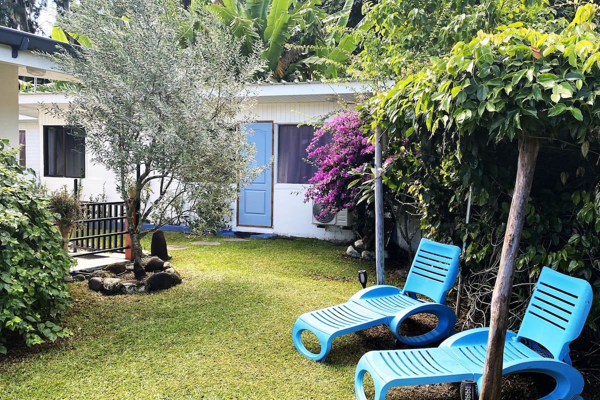 A well-maintained garden with a lawn, two blue lounge chairs, a small tree, and a house with a blue door and air conditioning unit.