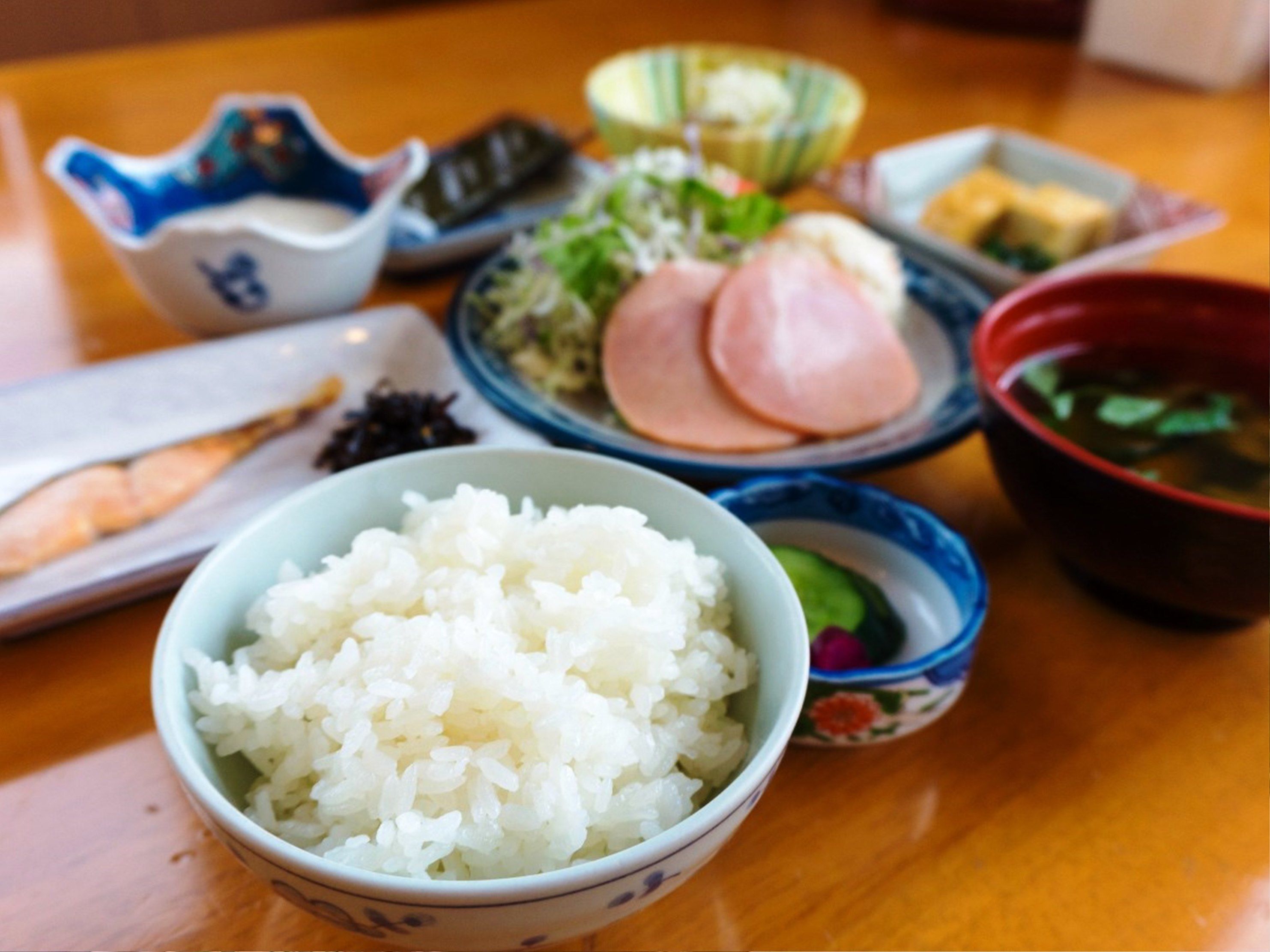 Una mesa con un tazón de arroz blanco, un plato de jamón en rodajas, un tazón pequeño de sopa y una taza con un diseño azul.