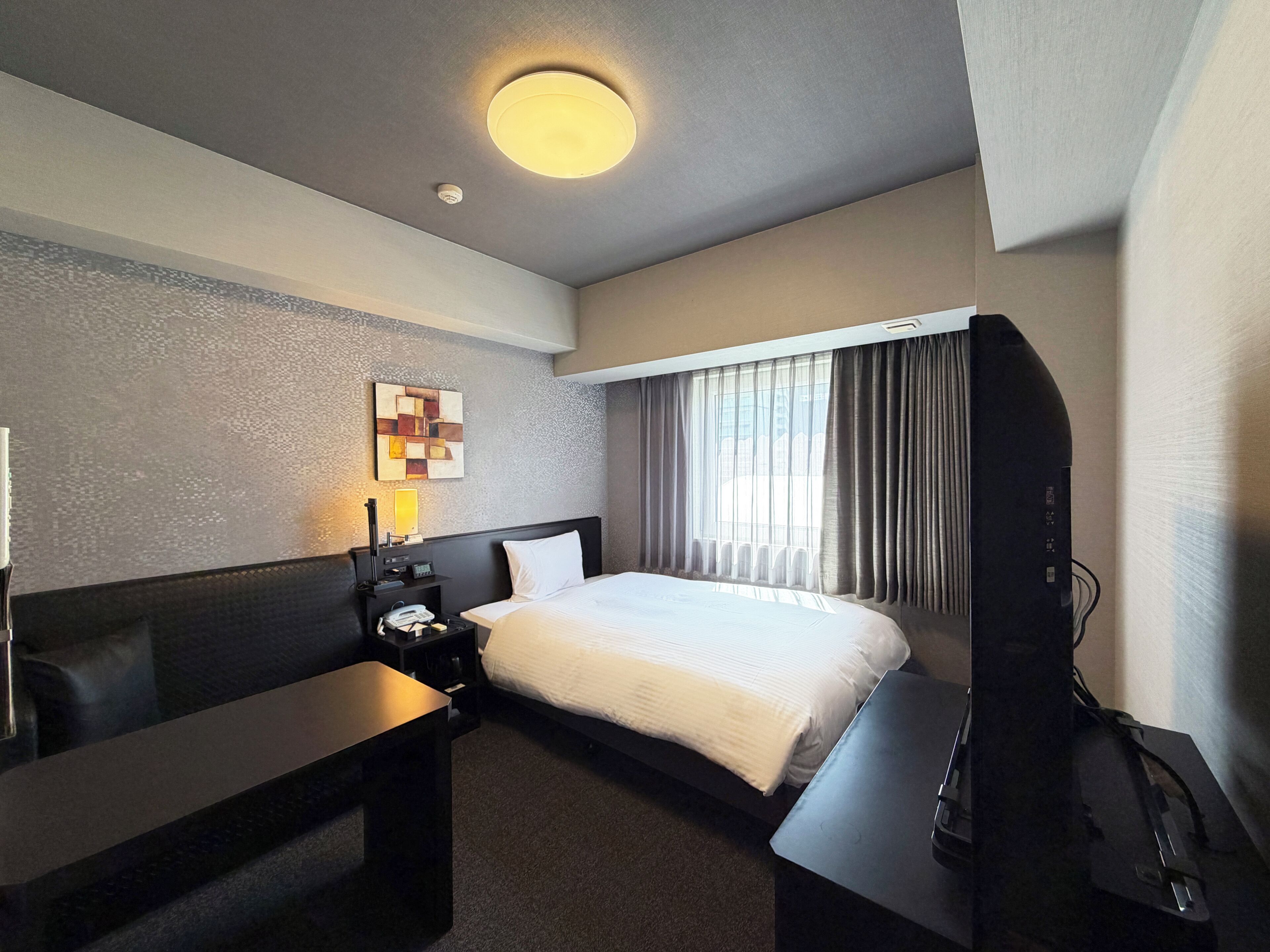 A hotel room with a bed, a desk, a TV, a lamp, and a wall-mounted artwork.