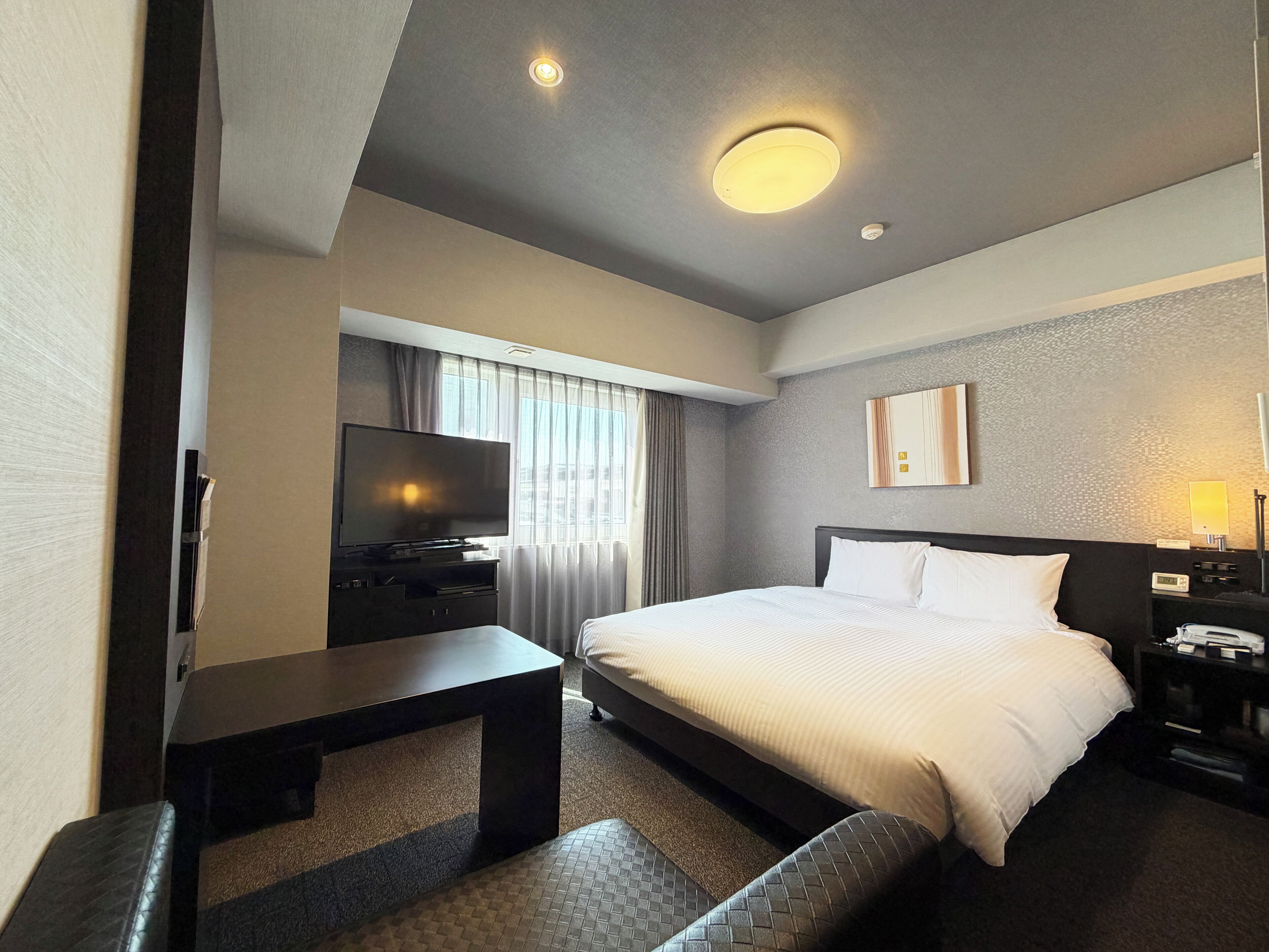 Quarto de hotel com cama, televisão, secretária e mesa de cabeceira.