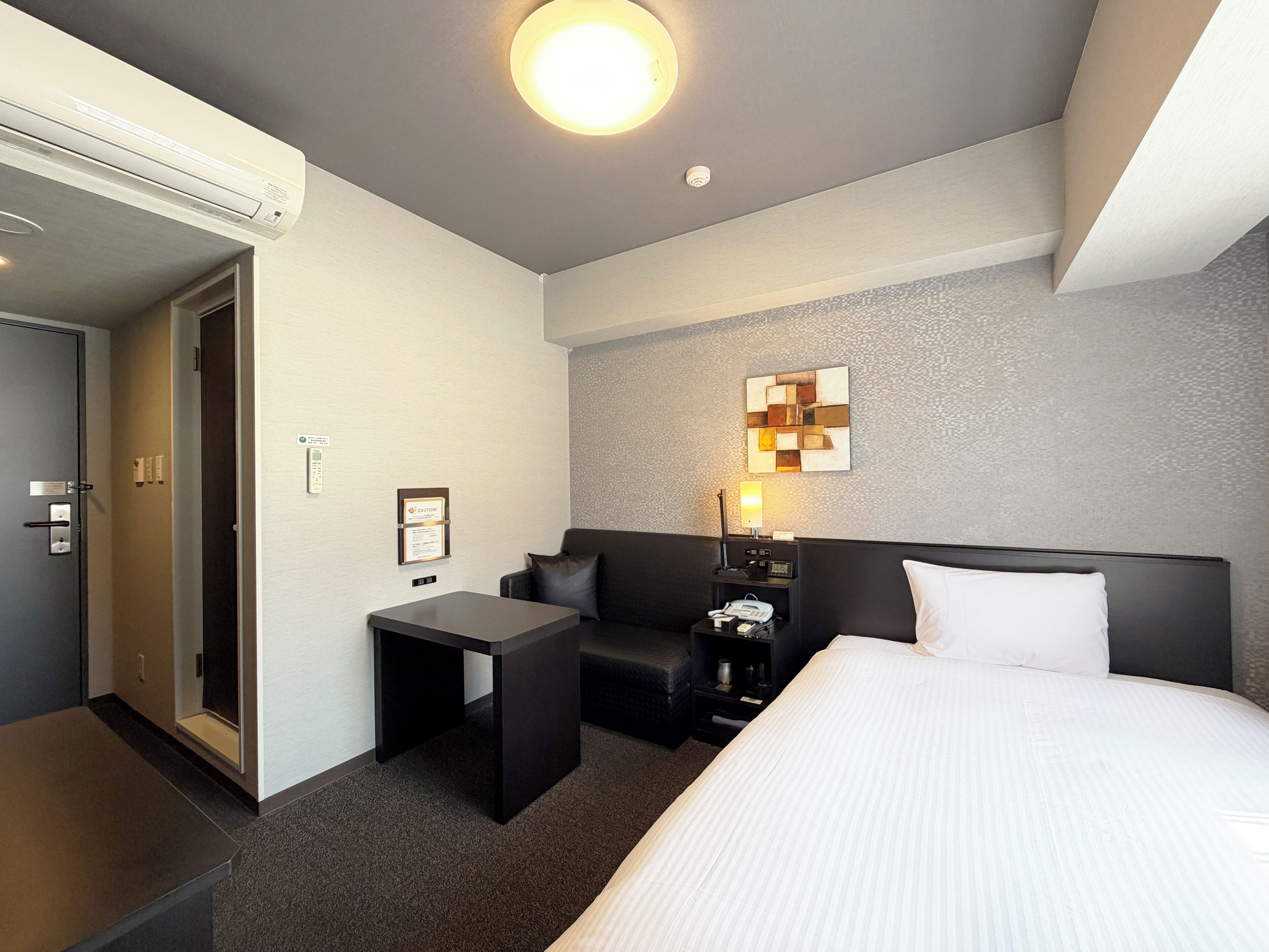 A hotel room with a bed, a desk, a sofa, a nightstand, and a wall-mounted artwork.