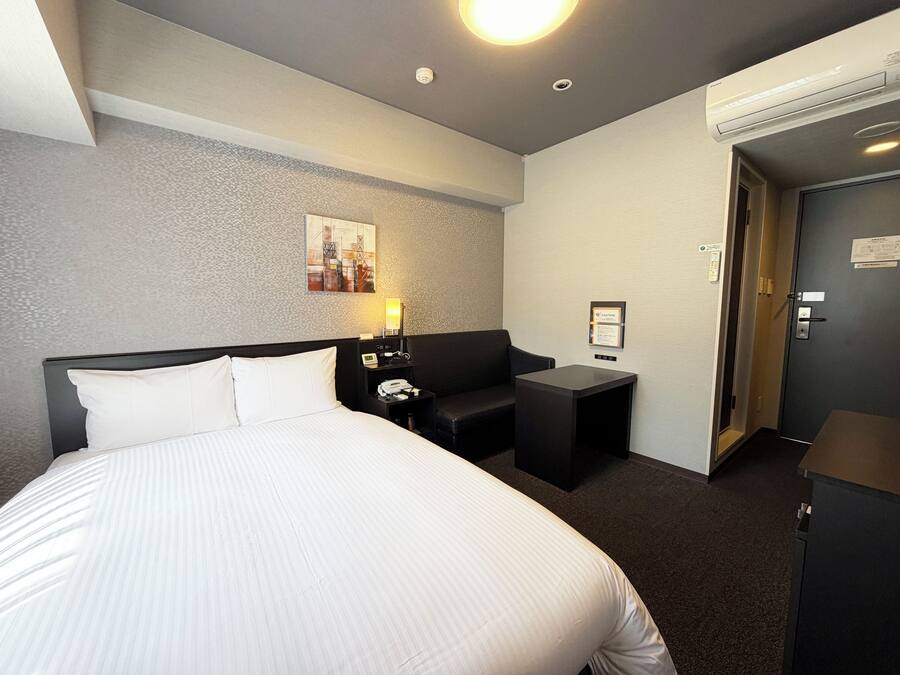 Et hotelværelse med en stor seng, et skrivebord, en stol, en lampe, et billede på væggen og en aircondition.