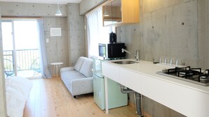 A compact kitchen with a built-in stove, a small refrigerator, and a sink. There is a dining table and a sofa in the adjacent living area. - Casa Botanika (Yomitan)