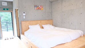 A bedroom with a wooden bed, white bedding, a concrete wall, a colorful artwork, and a door leading outside. - Casa Botanika (Yomitan)
