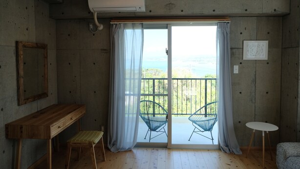 A room with a wooden bench, a small table, and a view of the sea through a balcony with chairs. - Casa Botanika (Yomitan)