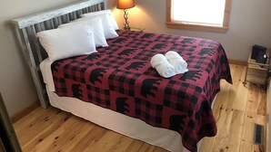 Una cama con una colcha a cuadros, almohadas blancas y toallas enrolladas. Hay una lámpara en una mesita de noche, una ventana con persianas y un tocador de madera con una computadora portátil.