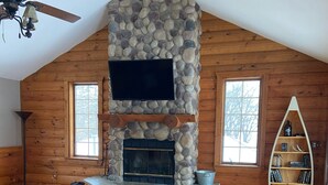 Interior acogedor de una cabaña de madera con chimenea de piedra, piso de madera, ventilador de techo y estantería.
