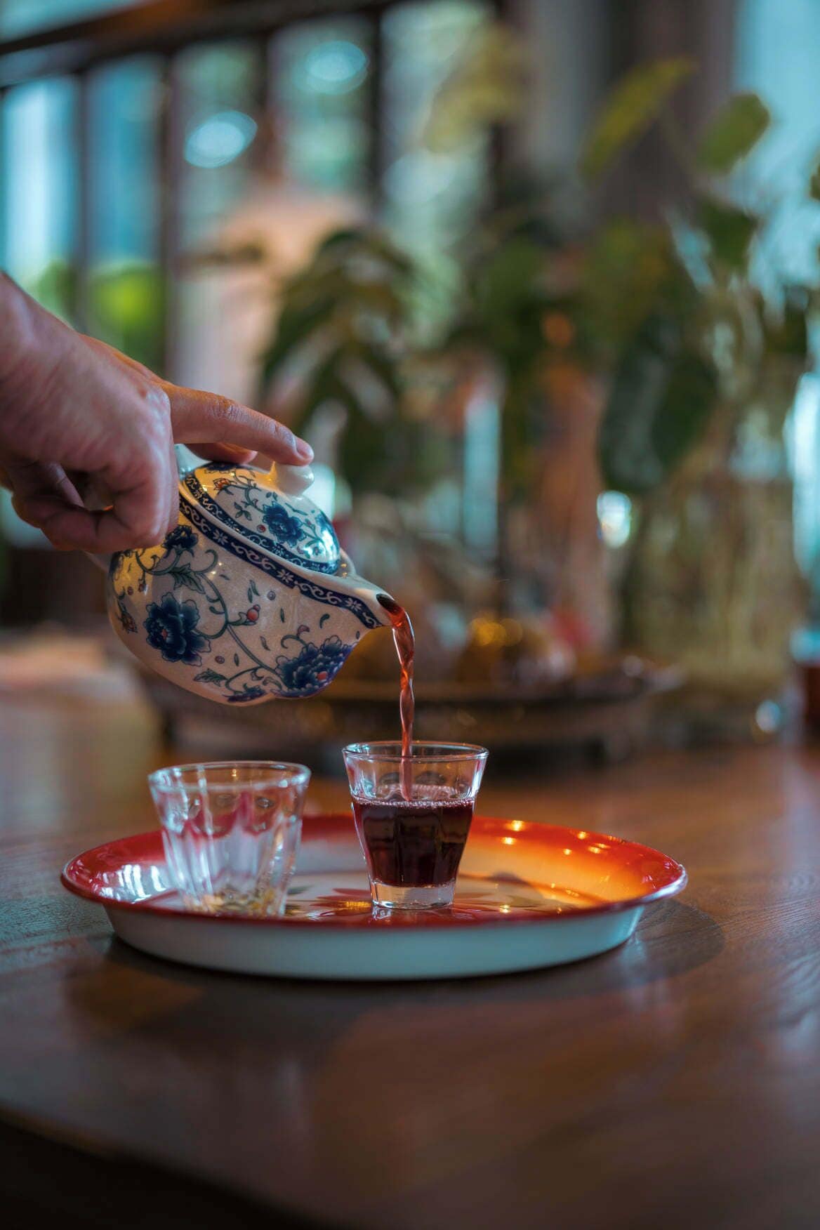 A hand pouring tea from a blue and white patterned teapot into a glass on a red tray.