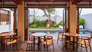 A spacious outdoor dining area with wooden tables and chairs, a view of a swimming pool, and lush greenery.