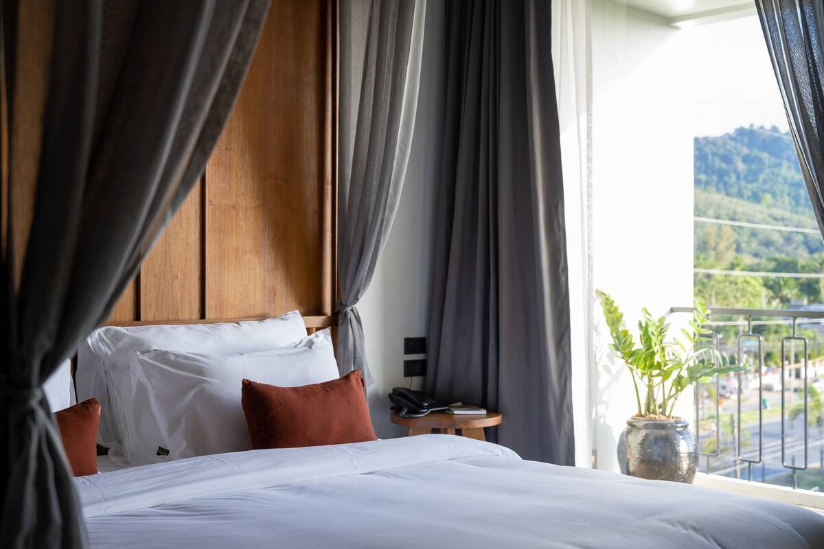 A hotel room with a large bed, a wooden headboard, a small table with a phone and a book, and a view of a mountain through a window.