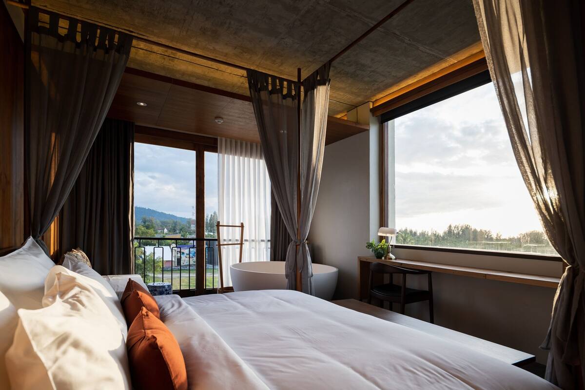 A bedroom with a large bed, a view of the outdoors, a small table with a lamp, and a chair.