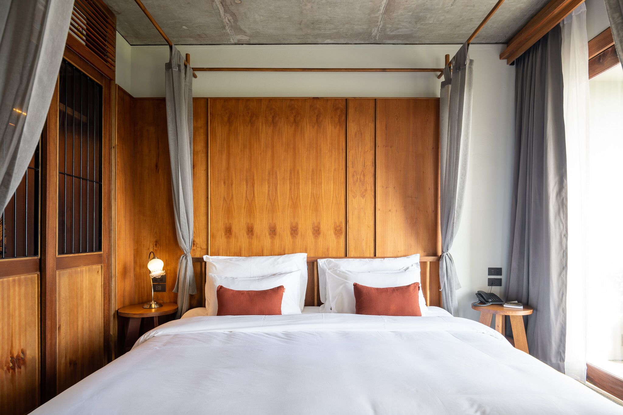 A hotel room with a wooden headboard, a bed with white linens, a bedside table with a lamp, and a small table with a phone and book.