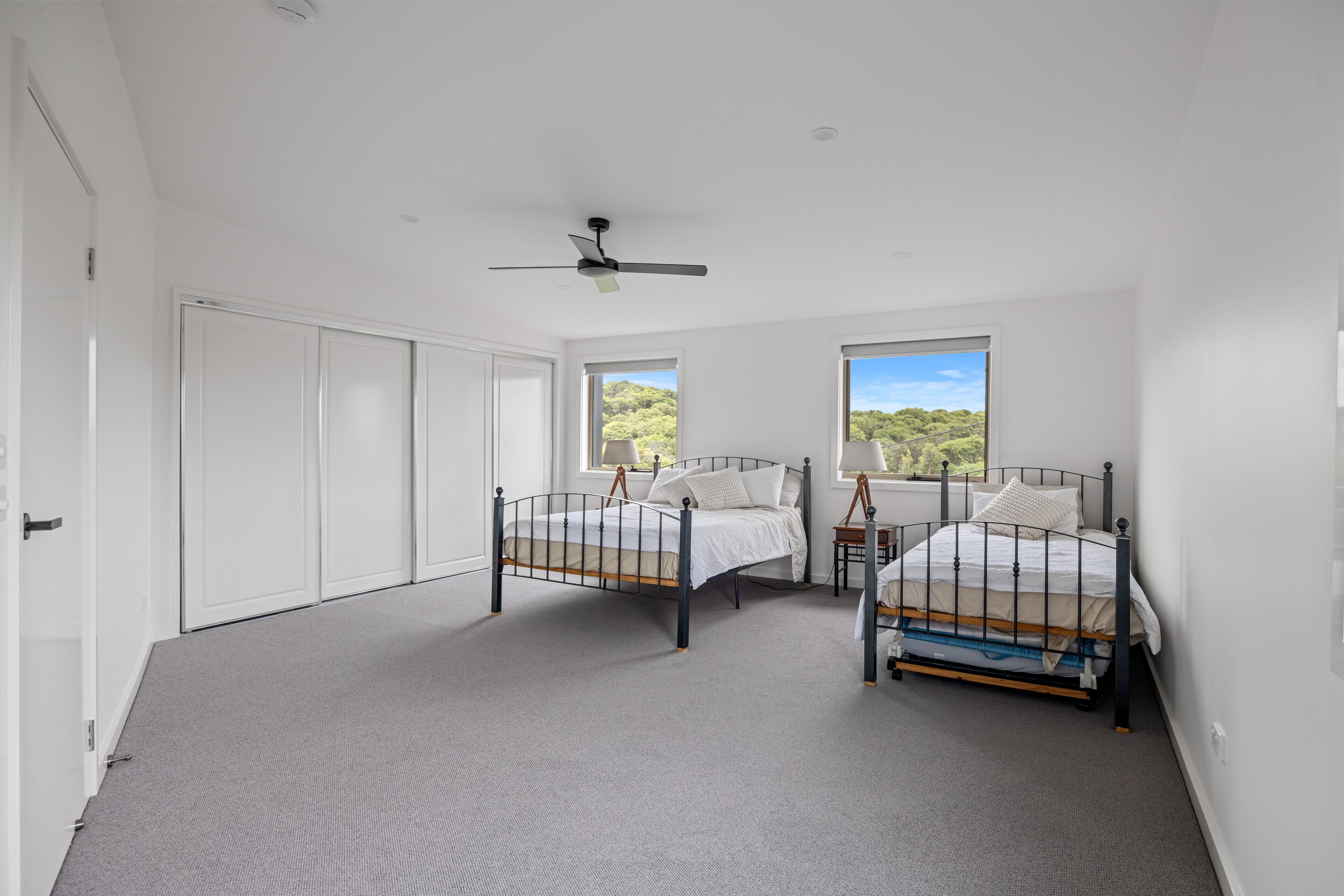 A bedroom with two beds, a ceiling fan, and large windows with a view of greenery.