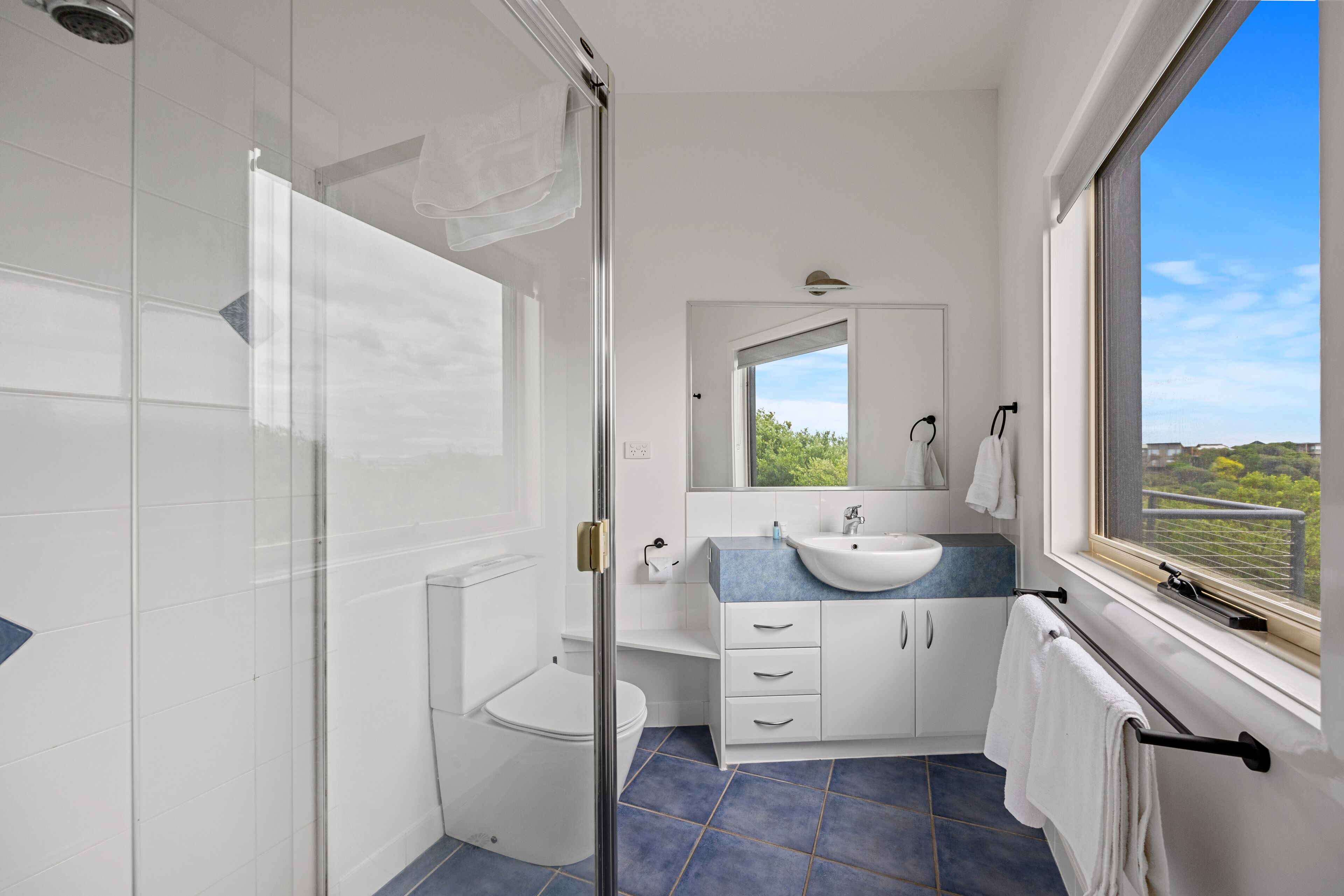 A modern bathroom with a glass shower enclosure, a white toilet, a sink with a blue countertop, and a window offering a view of greenery.
