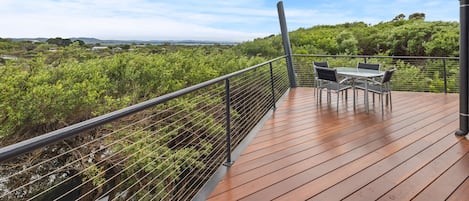 Une terrasse en bois avec une table et des chaises, offrant une vue sur un paysage verdoyant, parsemé d’arbres, et sur la mer au loin.