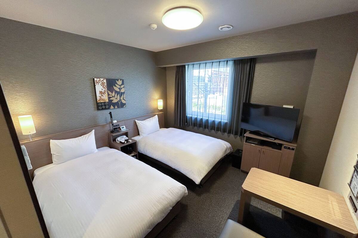 A hotel room with two beds, a desk, a TV, and a window with curtains.