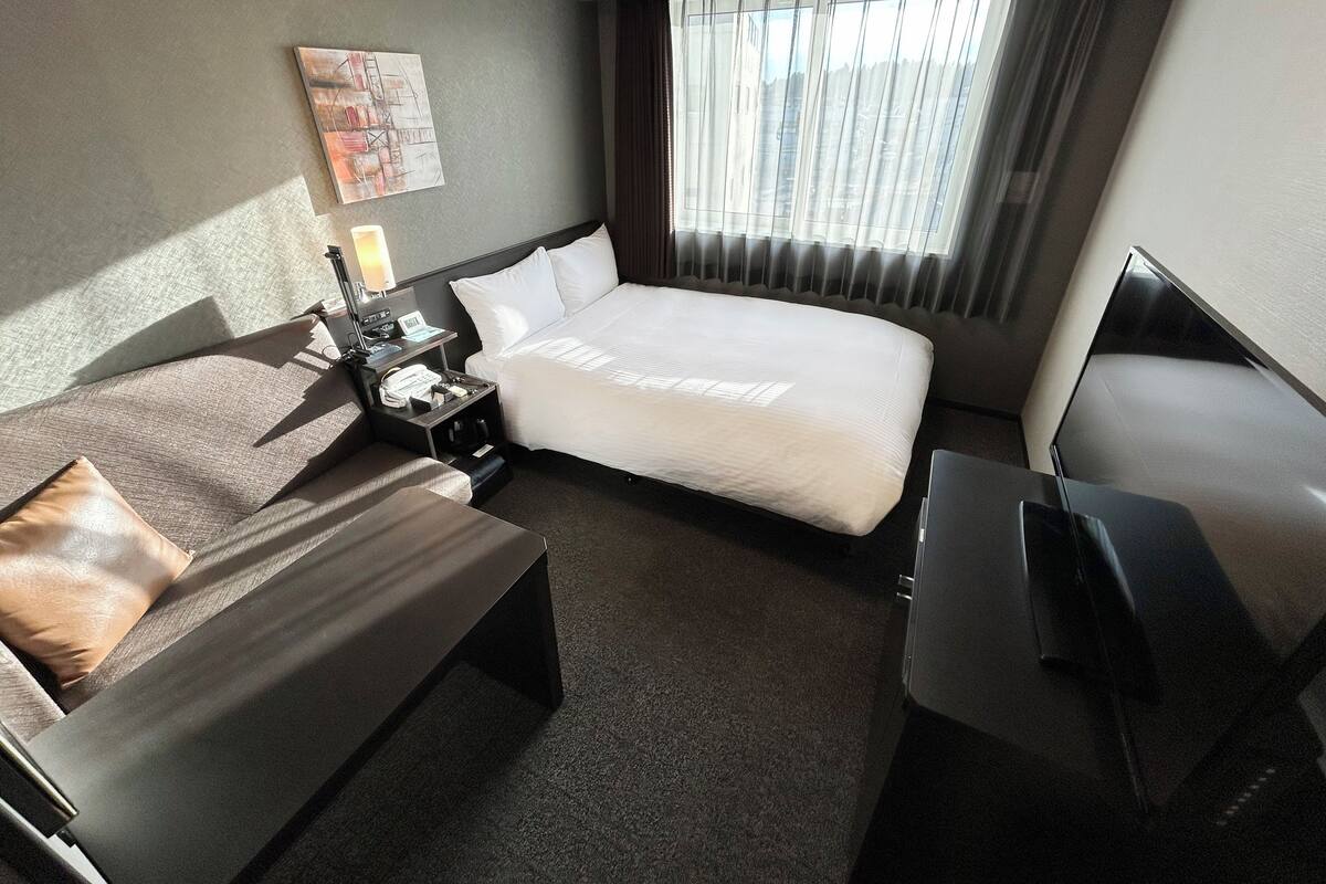 A modern hotel room with a large bed, a desk, a chair, a TV, and a window with curtains.