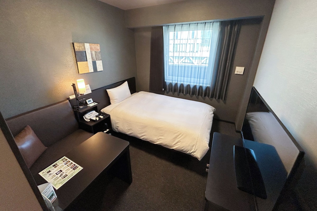 A hotel room with a bed, a desk, a chair, a nightstand, a lamp, and a window with curtains.