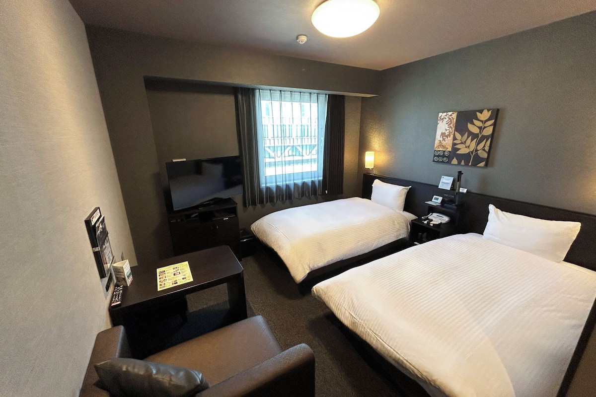 A hotel room with two beds, a desk with a flat-screen TV, a small seating area, and a window with curtains.