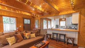 A cozy cabin interior with a wooden ceiling, a living area with a sofa and cushions, a kitchen with a bar stool, and a dining table.