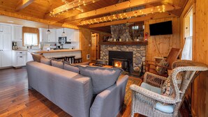 A cozy cabin interior with a stone fireplace, wooden ceiling beams, a gray sofa, wicker chairs, and a kitchen area.