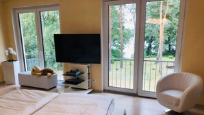 Un dormitorio con cama, televisión sobre un mueble, una silla y ventanas amplias con vista a árboles.