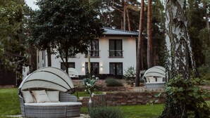 Una casa moderna con un muro de ladrillo y fachada blanca. En primer plano, dos sillones blancos de mimbre con cojines.