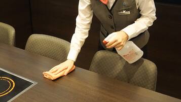A person in a white shirt and grey vest cleaning a wooden table with a cloth and a bottle of cleaning solution.
