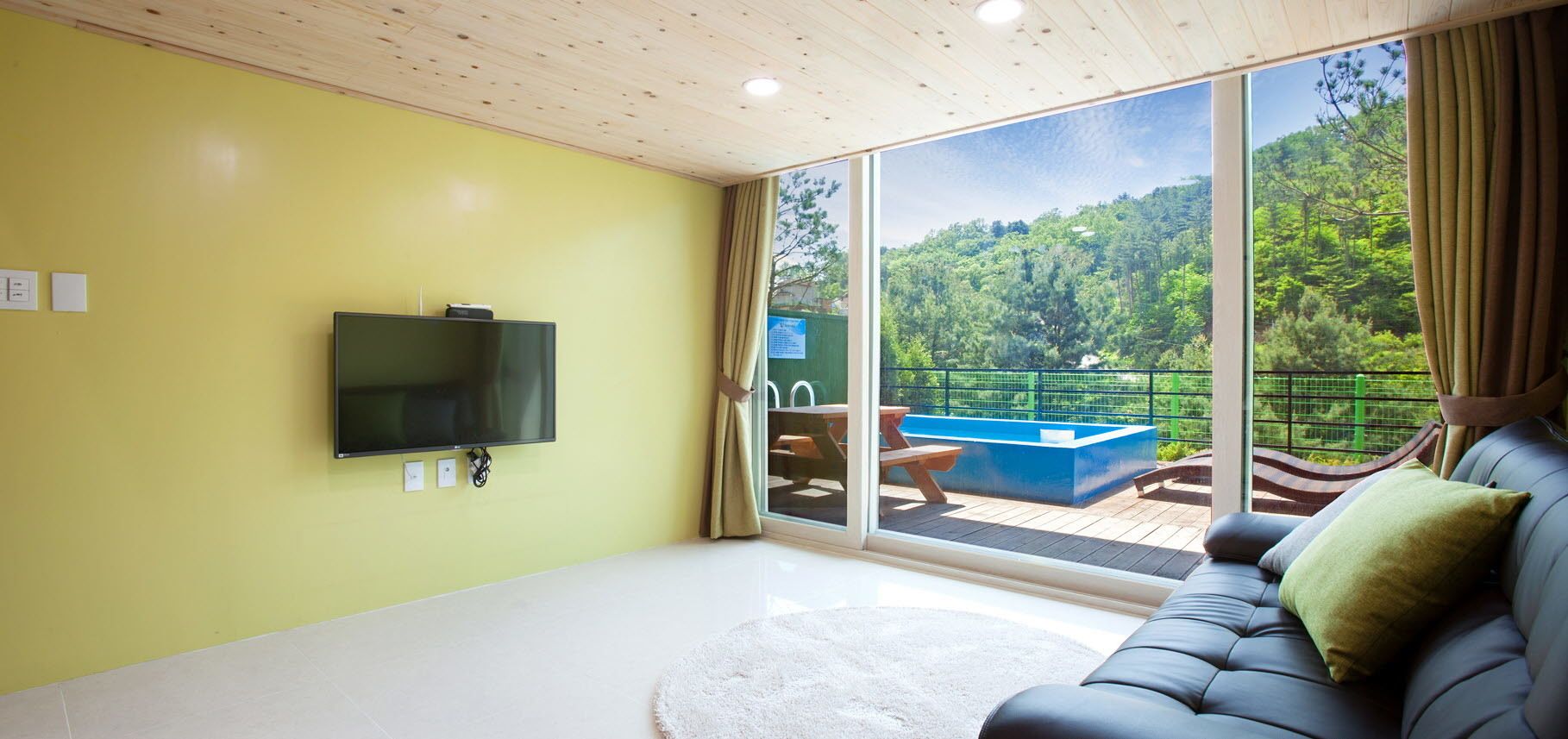 A room with a large TV, a sofa, and a sliding glass door leading to a balcony with a pool and outdoor furniture.