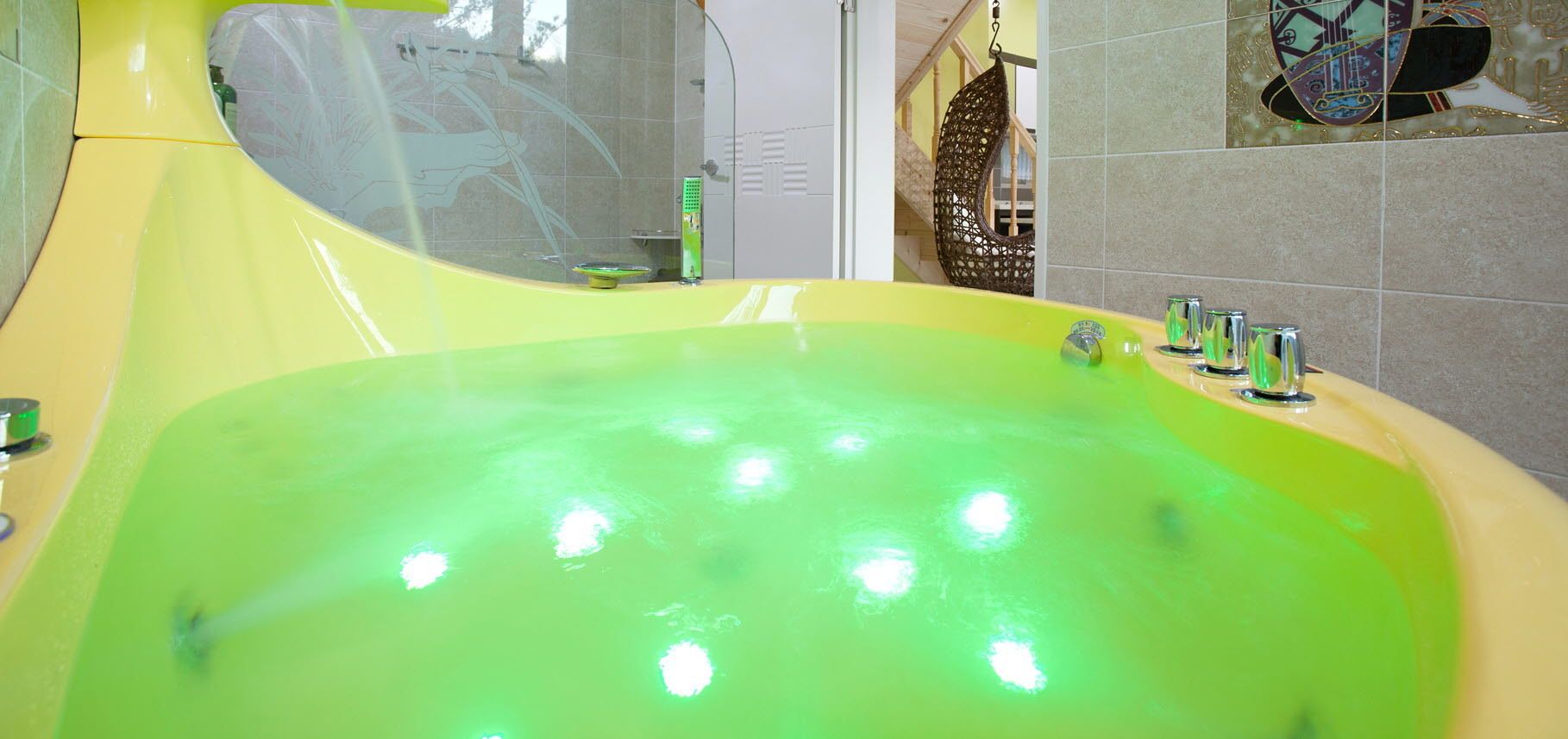 A yellow bathtub with a green interior, a glass shower enclosure, and a hanging chair visible in the background.