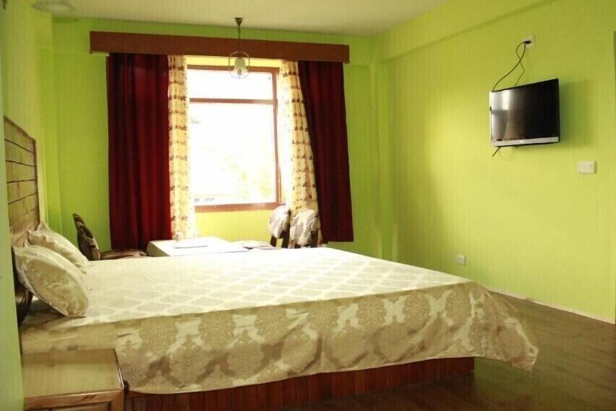Un dormitorio con cama, ventana con cortinas, televisión en la pared y puerta que da a otra habitación.