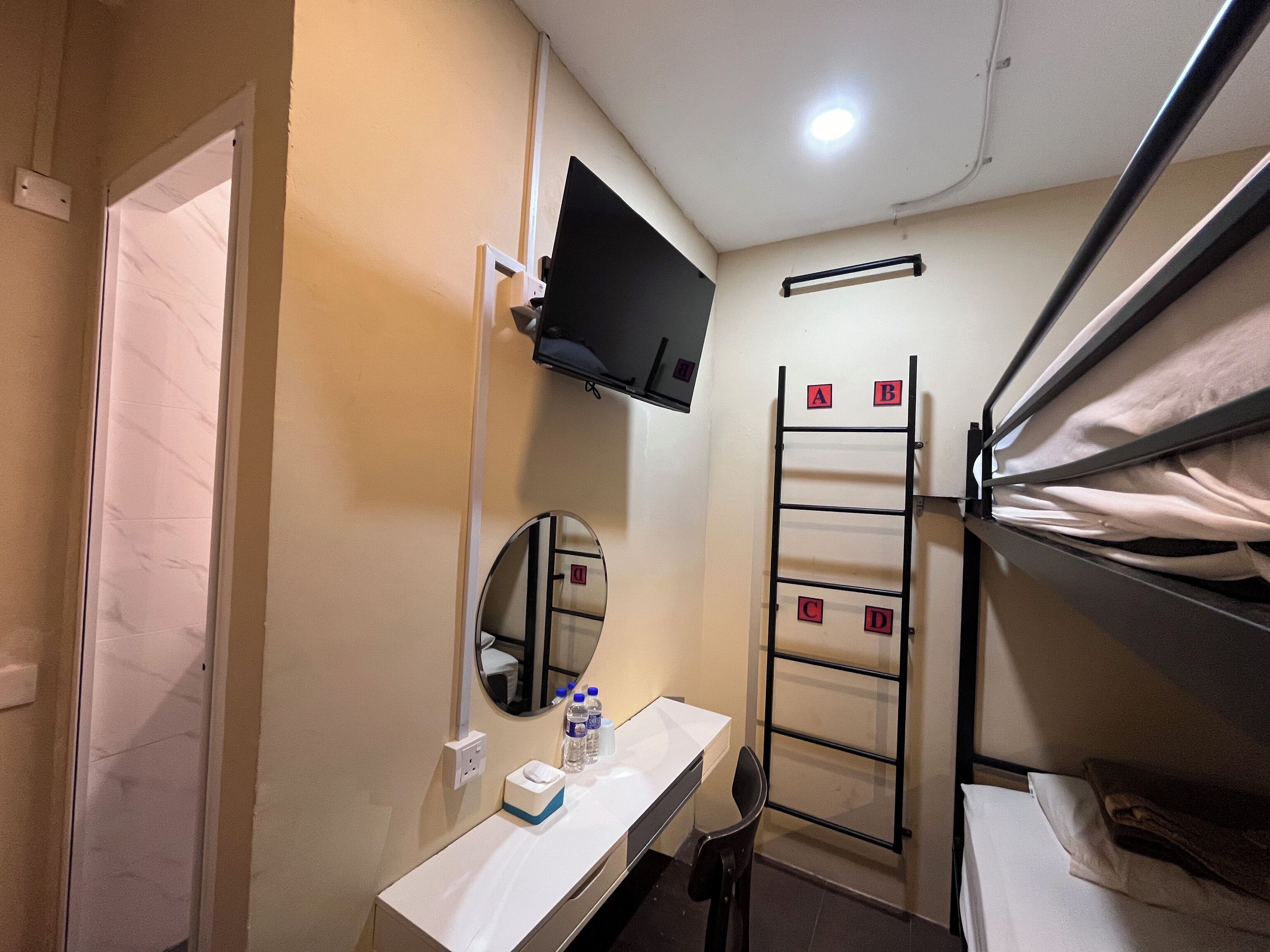 Een compacte hotelkamer met een stapelbed, een klein bureau met een spiegel, een aan de muur bevestigde televisie en een ladder om het bovenste bed te bereiken.