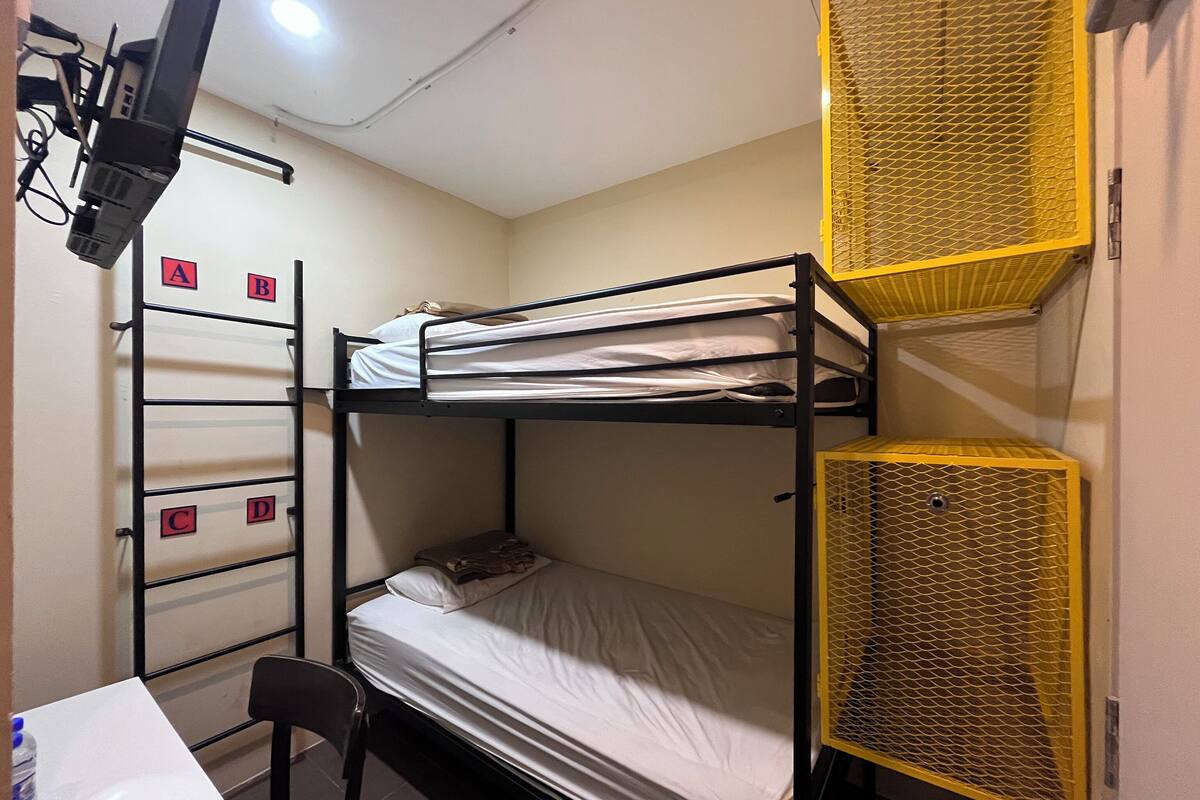 Une chambre avec un lit superposé, un bureau et une chaise, une étagère avec des compartiments étiquetés et un système de ventilation.