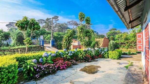 Un vialetto nel giardino conduce a un edificio con una porta rossa, circondato da vegetazione lussureggiante e piante fiorite.