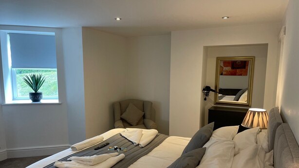 A modern bedroom with a large bed, a chair, a desk, and a window with a plant. - Beach House Courtyard 1 bed (Southport)