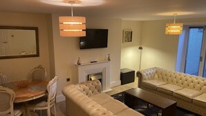 A modern living room with a beige sofa, a dining table with chairs, a fireplace, a flat-screen TV mounted on the wall, and a ceiling light fixture. - Beach House Courtyard 1 bed (Southport)