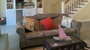 Ein Wohnzimmer mit einem braunen Sofa, einem Glastisch und einem schwarz-weißen Teppich. Eine Treppe mit weißen Geländern und ein Deckenventilator sind ebenfalls vorhanden.