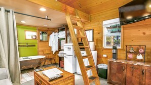 Innenansicht einer Holzhütte mit einer Holzleiter, einem Kühlschrank, einem Spülbecken, einem Tisch mit Fernbedienung und einem Schrank.