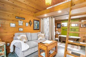 Ein gemütliches Zimmer mit Holzbalkendecke, einem grauen Sofa, einem Holztisch und einer Badewanne im Vintage-Stil im Hintergrund.