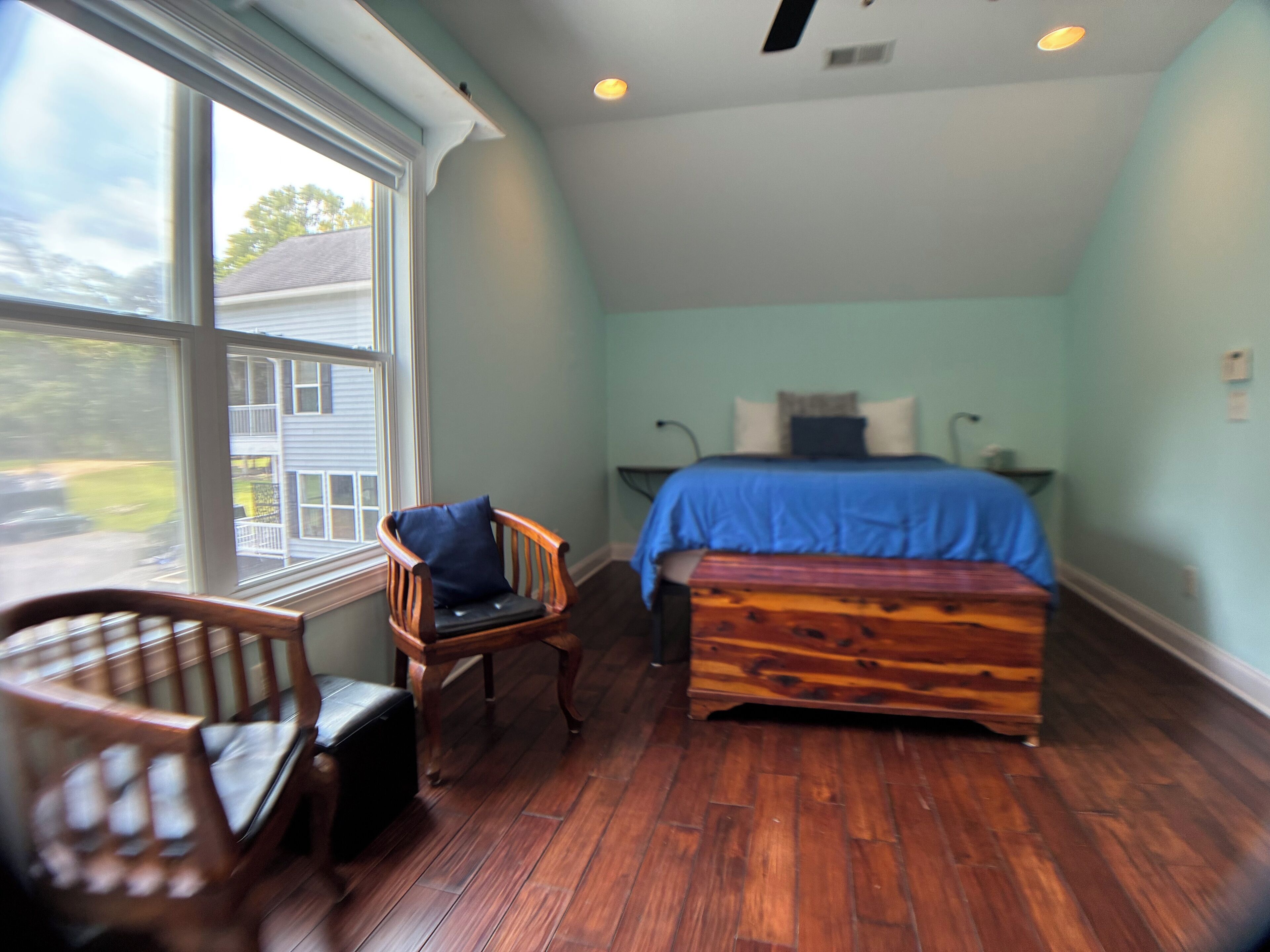 A bedroom with a bed, a chair, a bench, and a window with a view of a house.