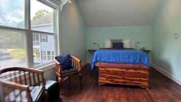 A bedroom with a bed, a chair, a bench, and a window with a view of a house.