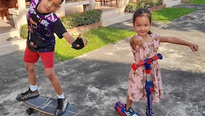 Two children on skateboards and a scooter in front of a building with a red roof and columns. - Sukthawee (Prachuap Khiri Khan)